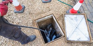 A technician feeds a drain rod into an open inspection chamber beside traffic cones, part of a job that factors into drain unblocking cost.